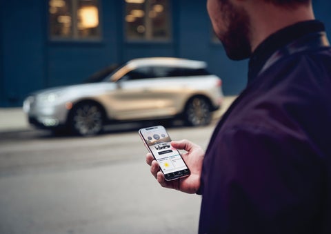 A man is holding his phone looking at the Lincoln Way™ App and we see his 2022 Lincoln Corsair SUV in the background against a blue brick wall | Bedford Lincoln PA in Bedford PA