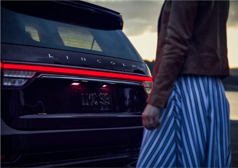 A person is shown near the rear of a 2023 Lincoln Aviator® SUV as the Lincoln Embrace illuminates the rear lights | Bedford Lincoln PA in Bedford PA