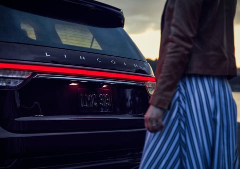 A person is shown near the rear of a 2024 Lincoln Aviator® SUV as the Lincoln Embrace illuminates the rear lights | Bedford Lincoln PA in Bedford PA