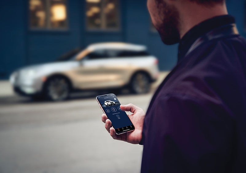 A person is shown interacting with a smartphone to connect to a Lincoln vehicle across the street. | Bedford Lincoln PA in Bedford PA